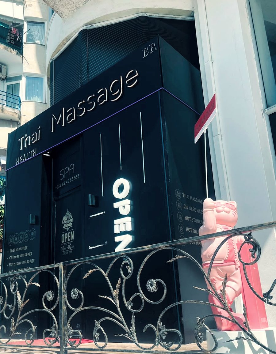 The front of Thai Health Massage on Rruga 40 Shenjtorët: a dark charcoal facade with Thai Massage Health in raised lettering, a lit OPEN sign, and a pink lucky cat beside the door.